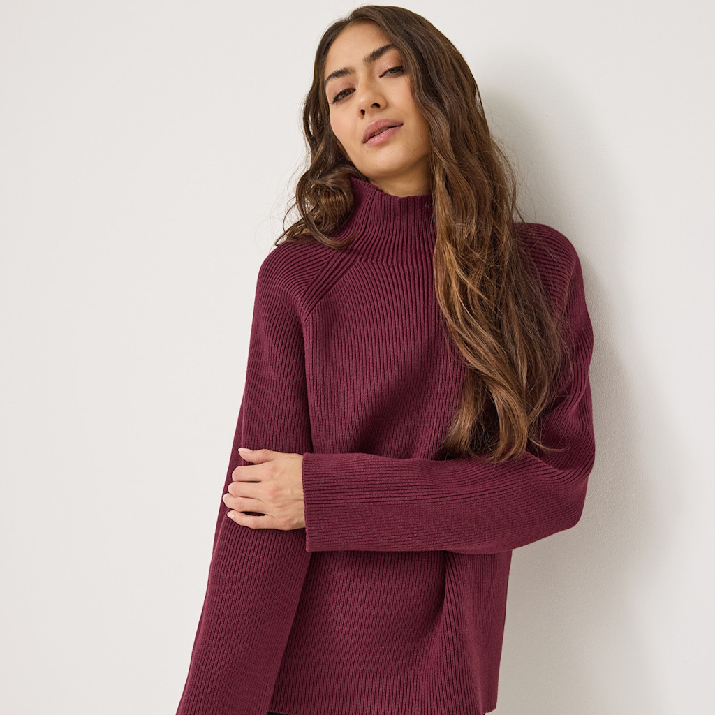 Modèle en pull-over tricoté bordeaux à col roulé posant sur fond blanc, longs cheveux bruns ondulés tombant sur le côté.