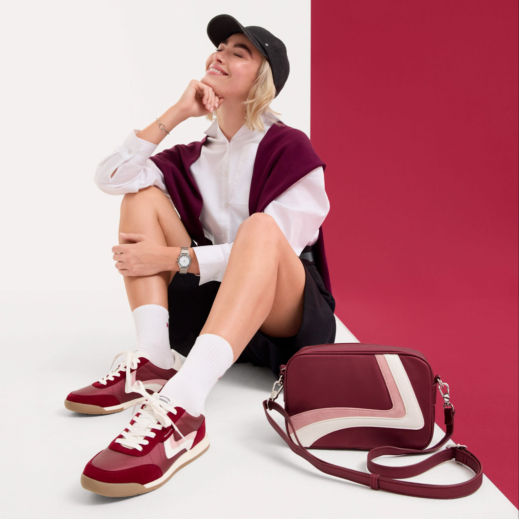 Modna stylizacja z bordowymi sneakersami i pasującą torebką, sportowo-elegancki wygląd z czapką z daszkiem na czerwonym tle.