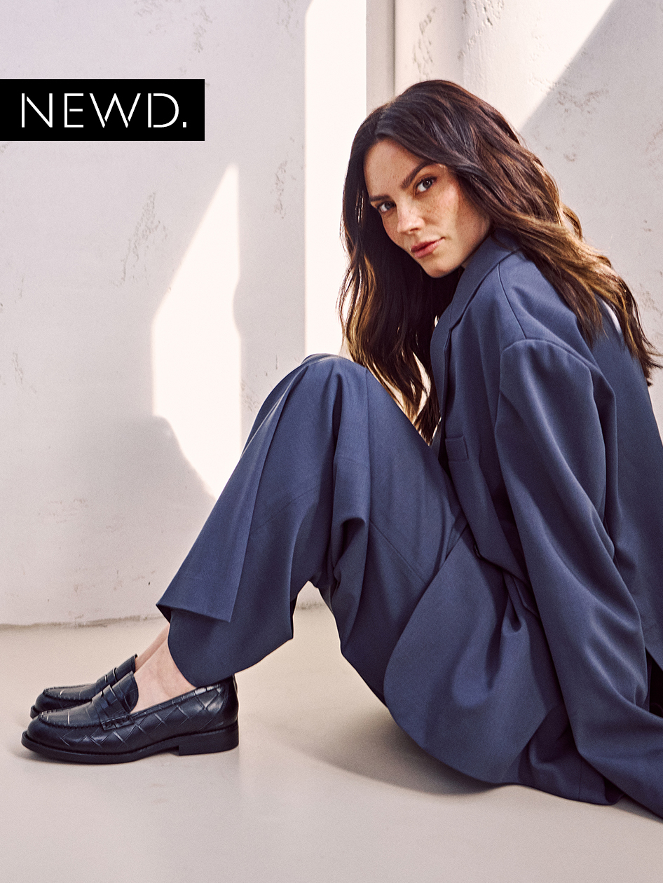 Model in elegant marineblauw broekpak zittend op de vloer, met gevlochten zwarte loafers. NEWD-logo op de foto.