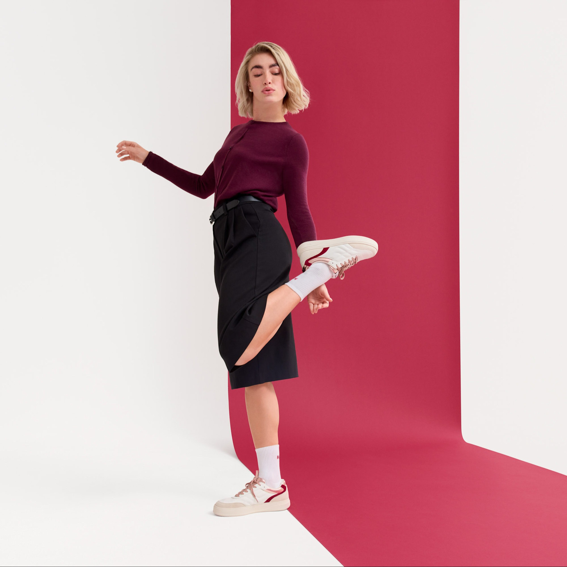 Modern outfit med vinröd tröja och svart kjol, dynamisk pose mot en rosa bakgrund, vita sneakers och strumpor.