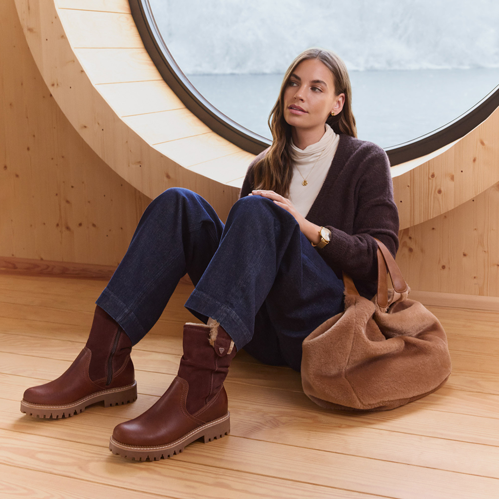 Junge Frau in Rollkragenpullover und Jeans sitzt entspannt in einem modernen Holzraum mit rundem Fenster, trägt braune Winterstiefel