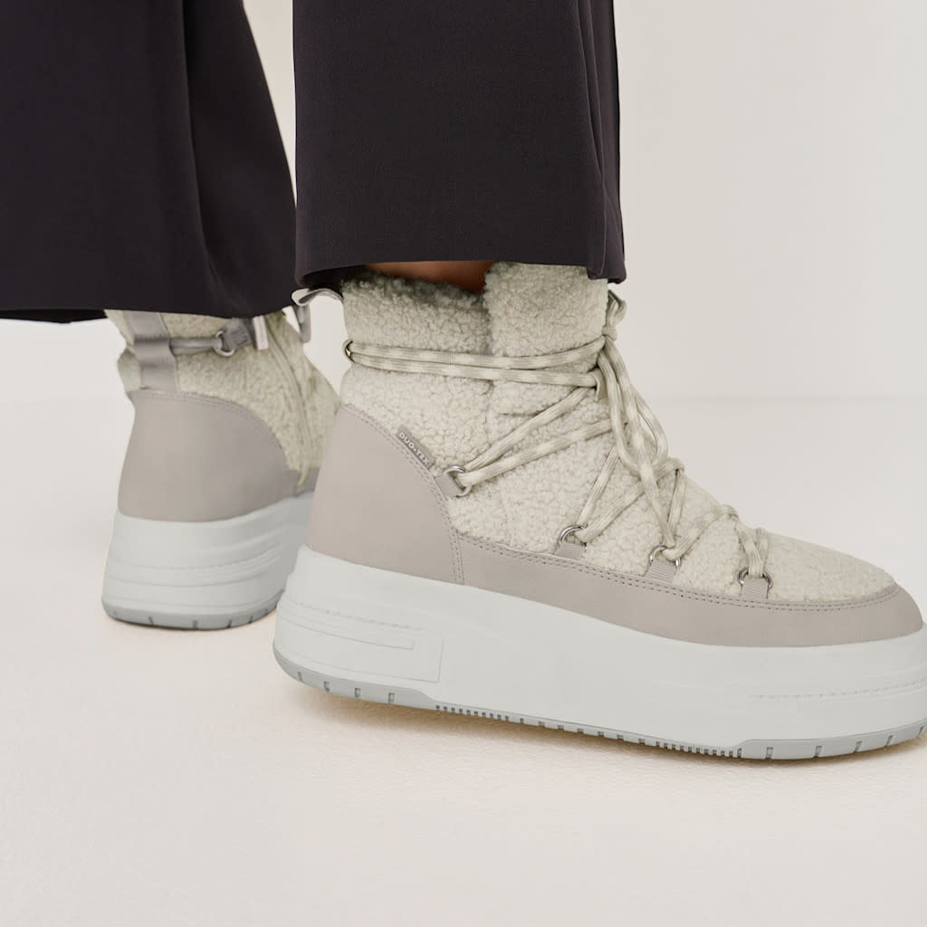 Hellgraue Plateau-Winterstiefel mit flauschigem Teddy-Besatz und weißer Sohle, getragen zu einer schwarzen Hose.