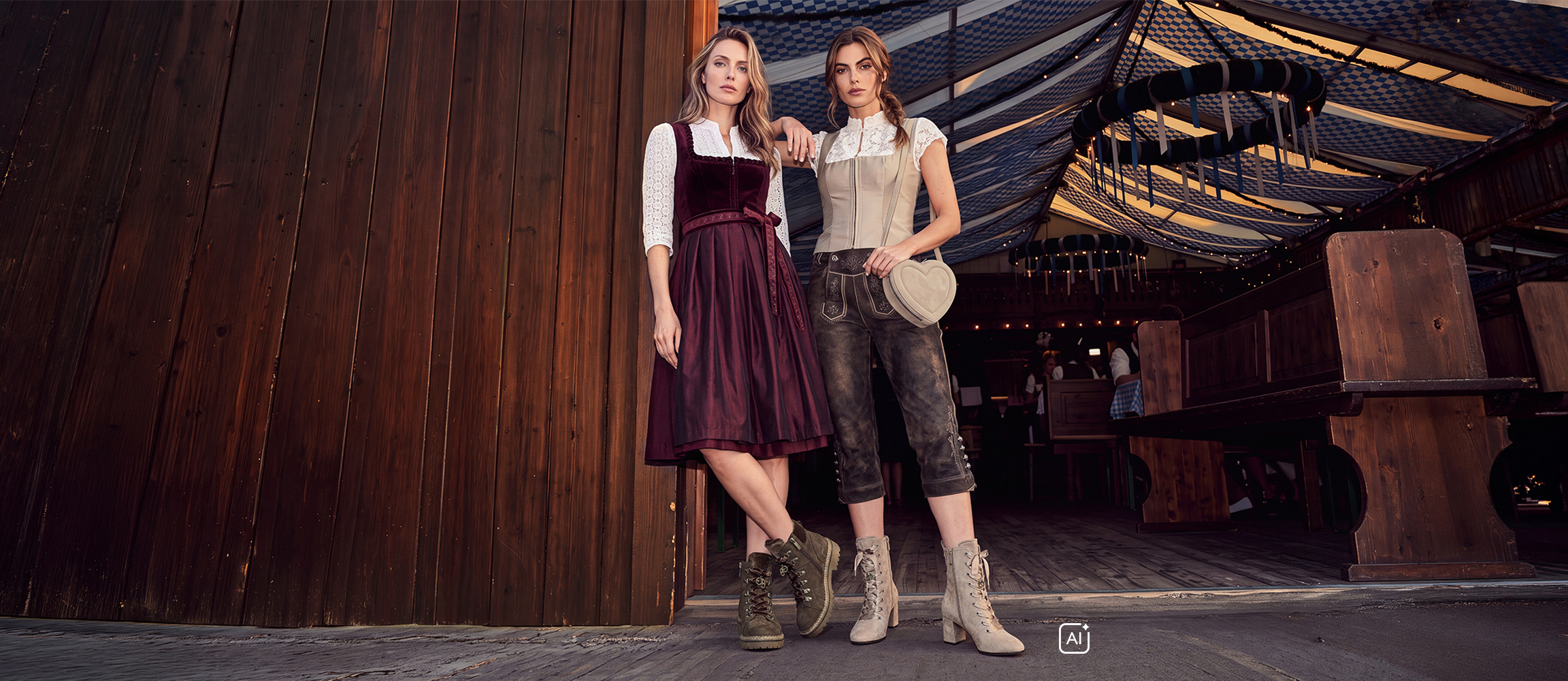 Zwei Frauen in Tracht: eine im weinroten Dirndl, eine in Lederhose mit beigem Mieder, vor einem Festzelt.