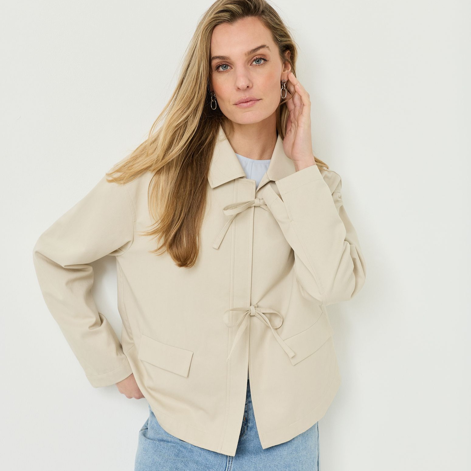 Vrouw met lang blond haar draagt een cr&egrave;mekleurige blouse met strikdetails en jeans voor een witte achtergrond.
