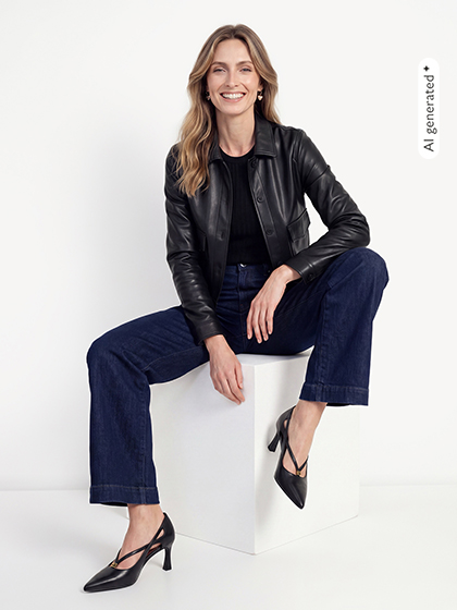 Glimlachende vrouw zit op een witte kubus, draagt een zwarte leren jas, donkere jeans en zwarte pumps.