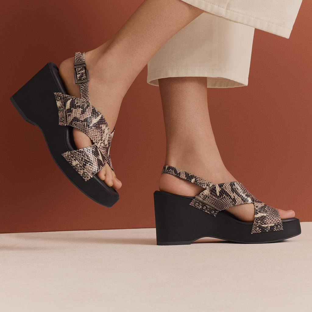 Moderne Sandalen mit Schlangenleder-Optik und schwarzer Plateausohle, pr&auml;sentiert vor terrakottafarbenem Hintergrund.