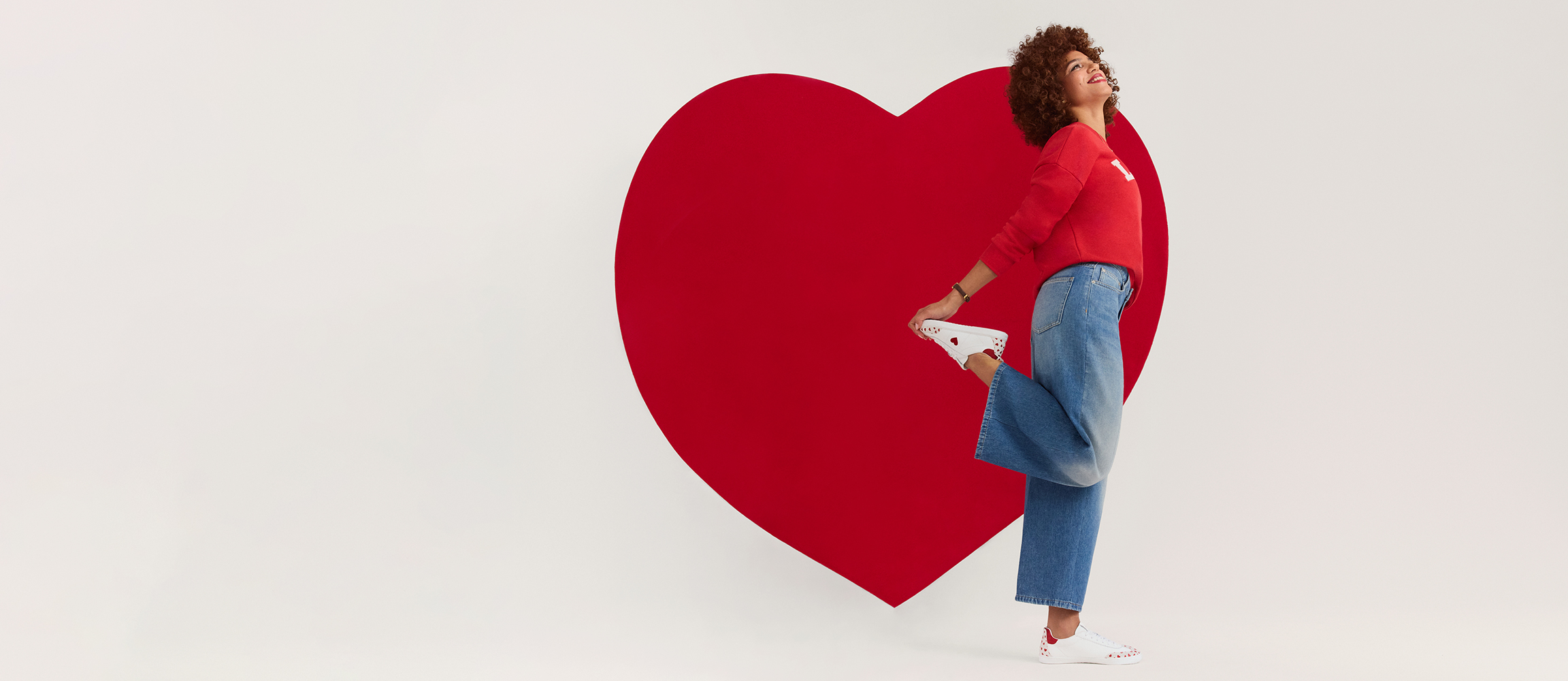 Person in a red sweater and jeans leans against a large red heart in front of a white background, looking upwards.