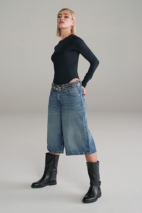 Frau in schwarzem Langarmshirt, weiten Jeans-Culottes, gemustertem G&uuml;rtel und schwarzen Lederstiefeln posiert vor grauem Hintergrund.