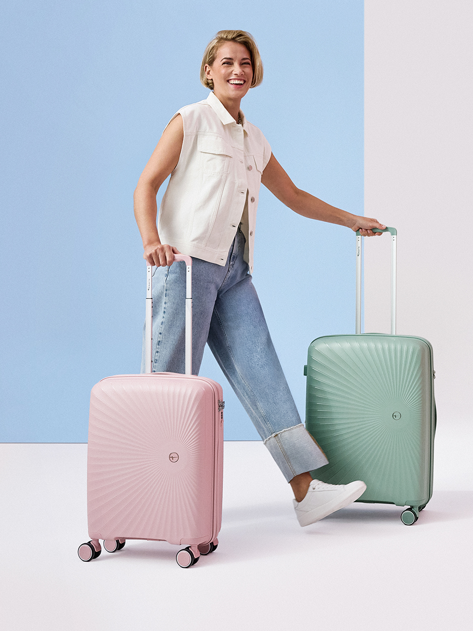 Donna sorridente con due trolley rigidi in rosa e verde menta, vestita con jeans e gilet bianco.