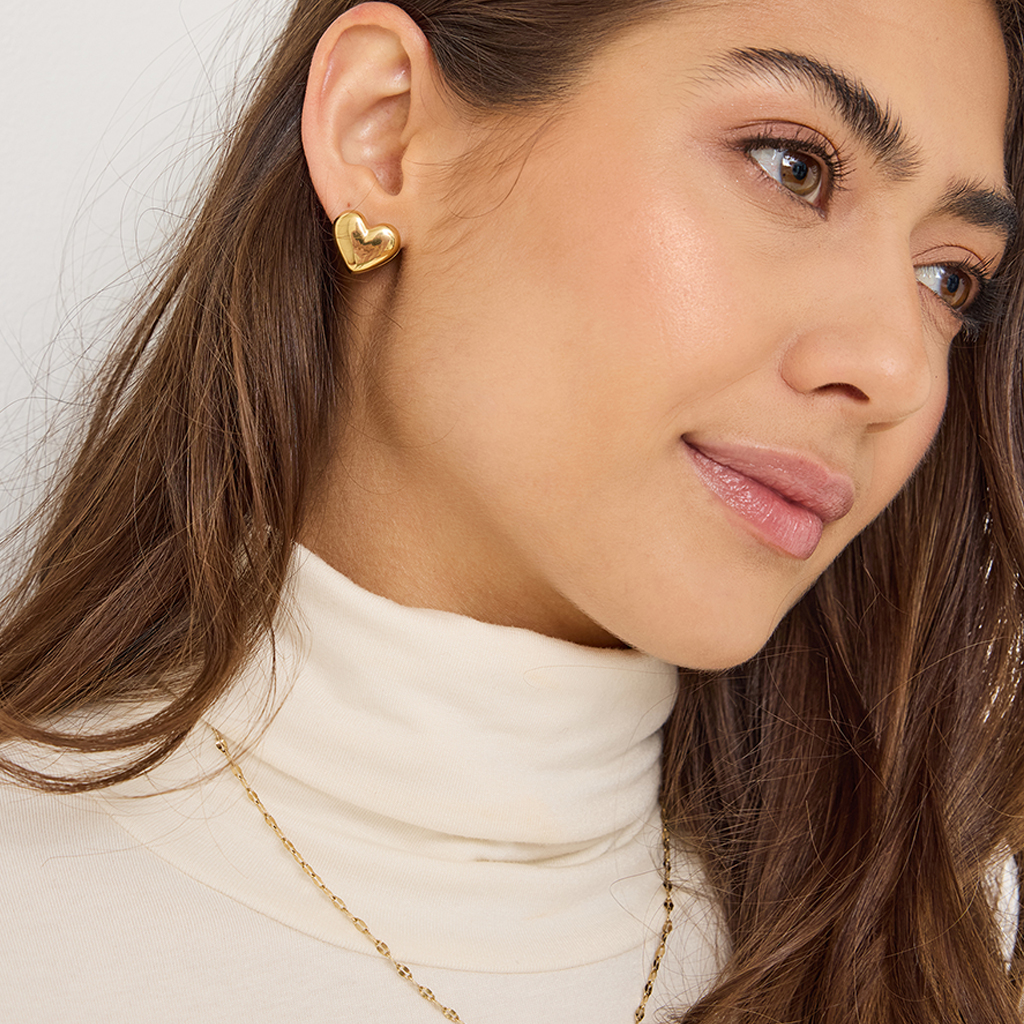 Close-up of a young woman in a white turtleneck sweater, with gold heart ear studs and natural makeup.