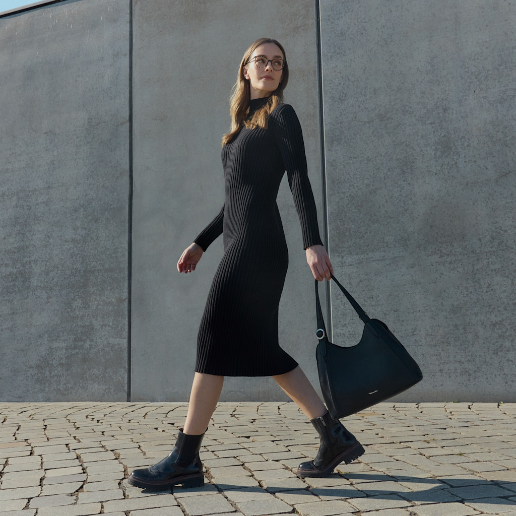 Elegant person in black knitted dress and boots walks with handbag in front of gray concrete wall on paved floor.