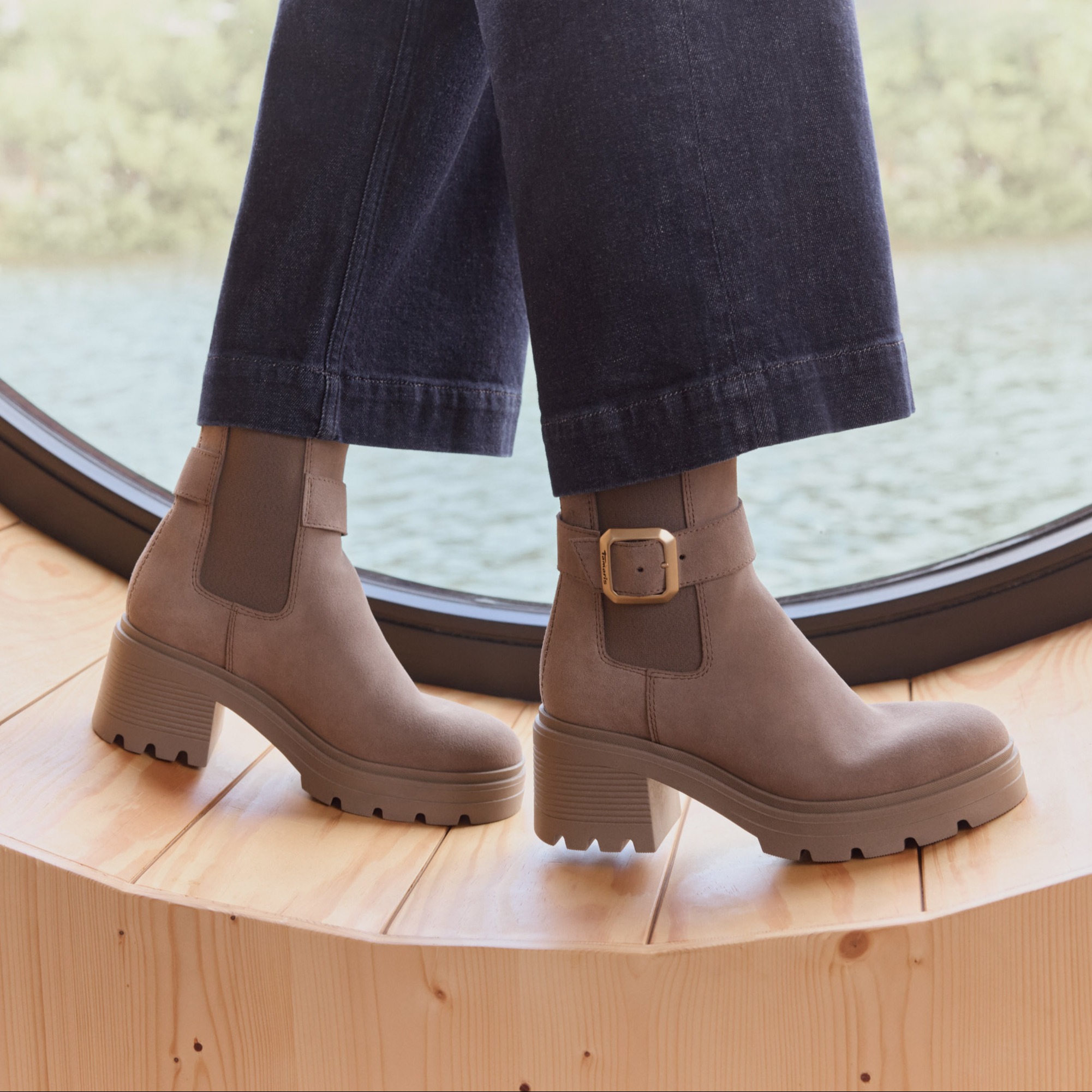 Stivaletti con tacco marrone e fibbia in look scamosciato, indossati con jeans scuri su un pavimento di legno chiaro.