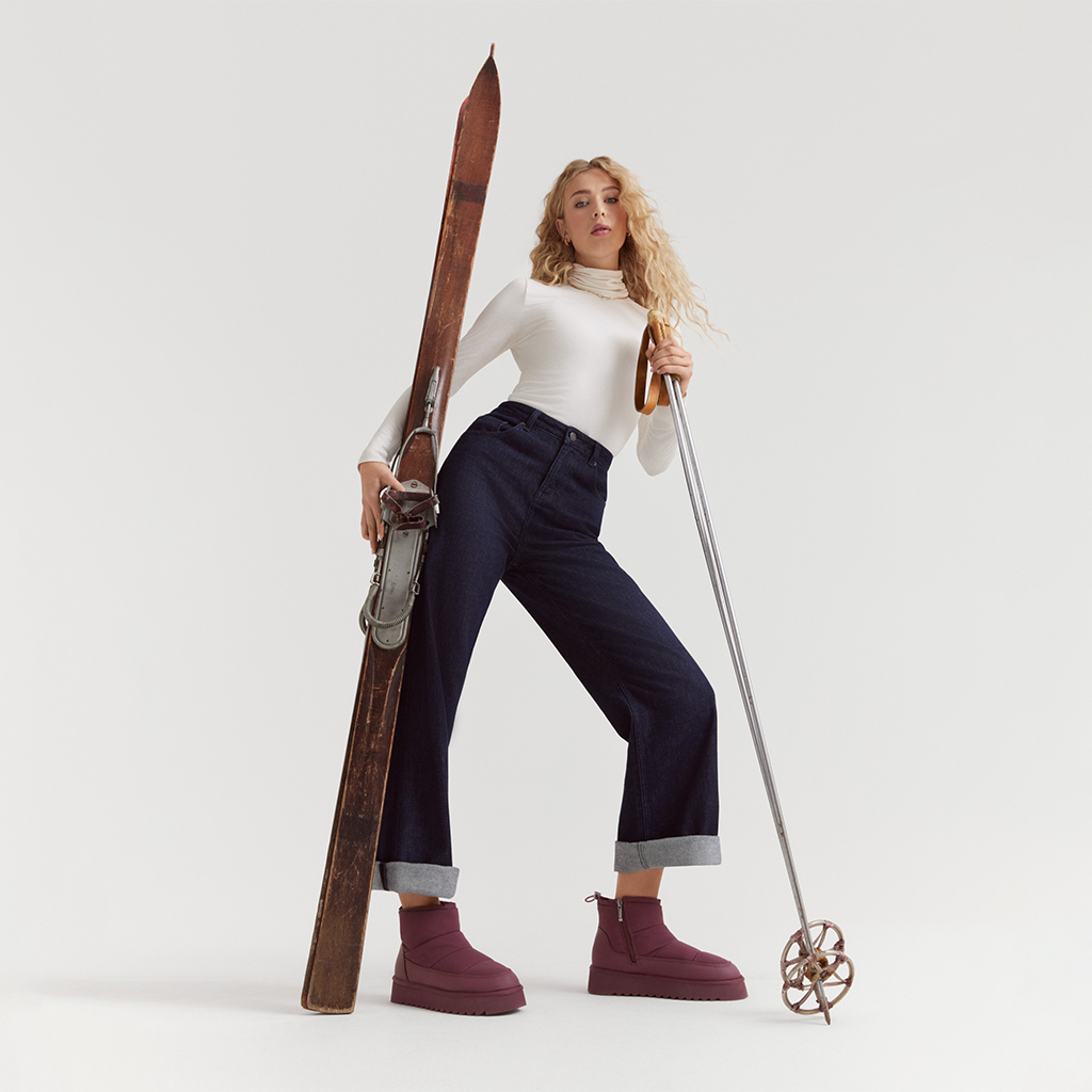 Modell i vit polotr&ouml;ja och jeans poserar med vintage skidutrustning, b&auml;r vinr&ouml;da vinterk&auml;ngor