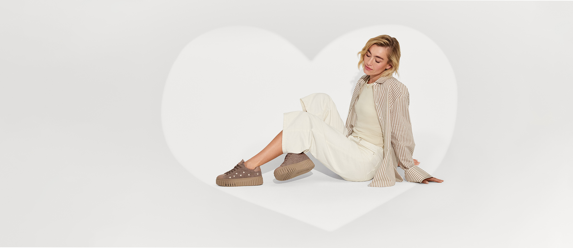 Woman sitting in front of a heart shape, wearing a cream-coloured outfit with a striped shirt and brown platform trainers on a white background.