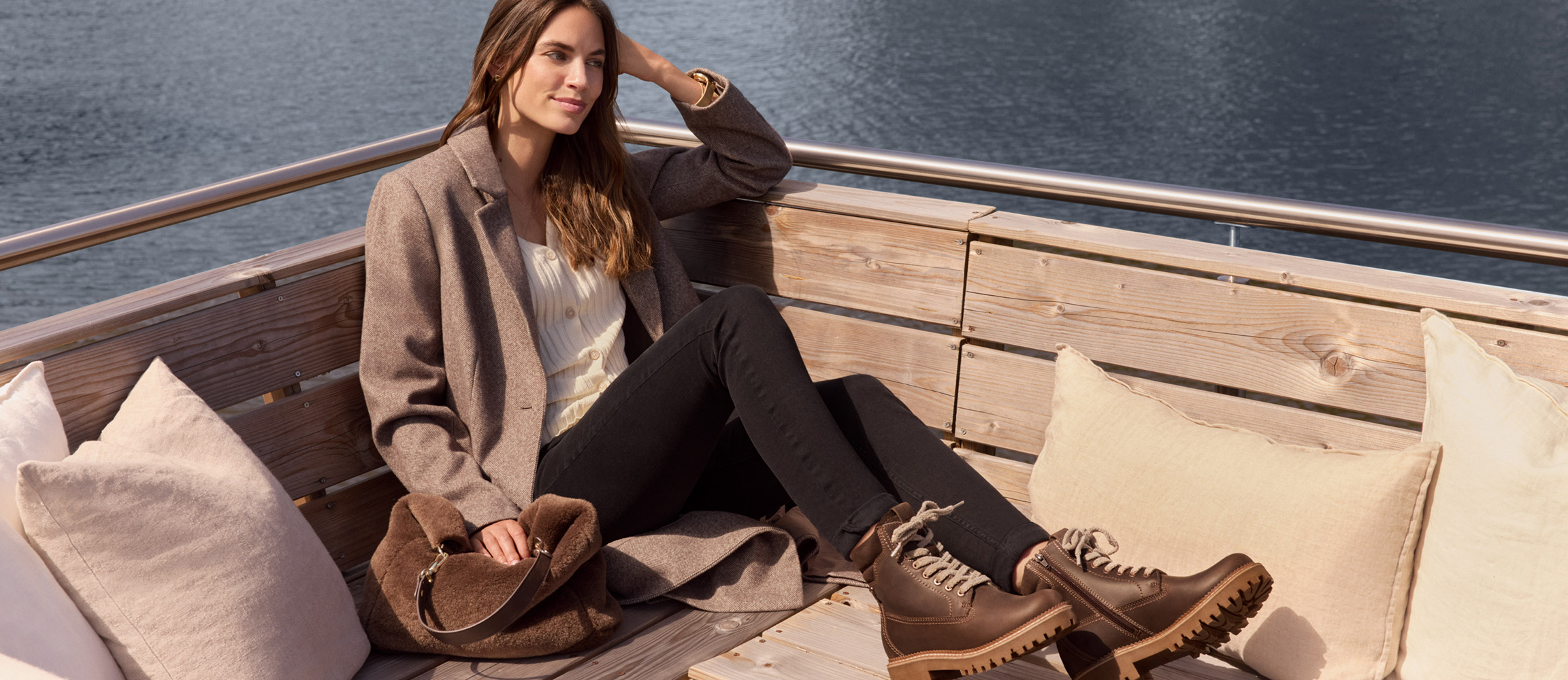 Modieuze herfstscène op een boot: Vrouw in beige jas en zwarte broek zit ontspannen op een houten bank met kussens en draagt bruine laarzen.