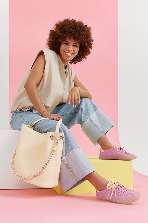 Modisches Outfit mit beiger Weste, Jeans und rosa Sneakers, dazu eine helle Handtasche vor pastellfarbenem Hintergrund