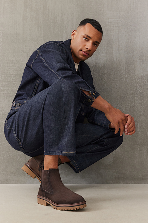 Mand i m&oslash;rkebl&aring;t denimoutfit b&aelig;rer brune Chelsea‑boots med profils&aring;l og sidder p&aring; hug foran gr&aring; baggrund.