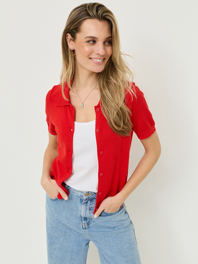 Glimlachende vrouw in een rood kortmouwig gebreid vest, wit topje en jeans tegen een lichte achtergrond, met lang blond haar en een halsketting.