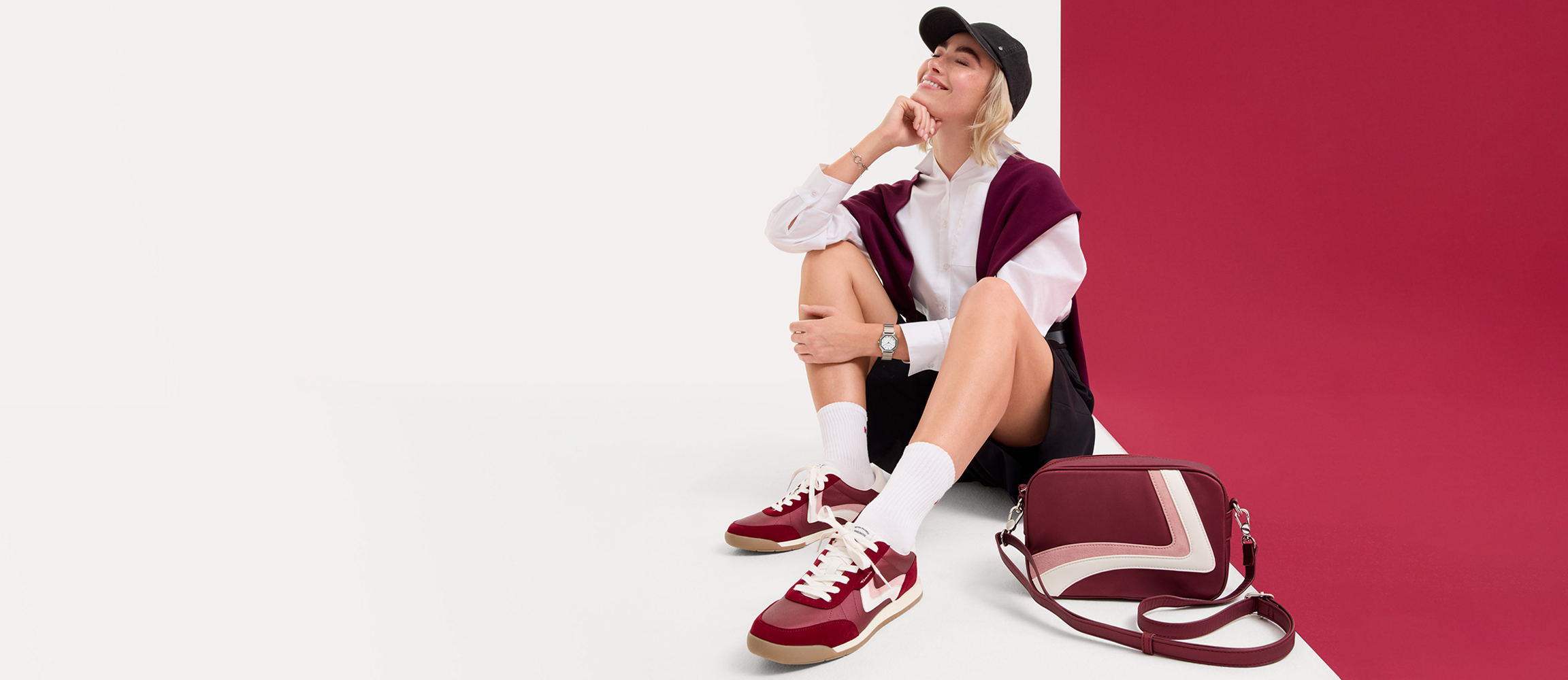 Personne habill&eacute;e de mani&egrave;re sportive, portant une casquette de baseball, un polo blanc et un pull bordeaux, avec des baskets bordeaux assorties sur fond rouge.