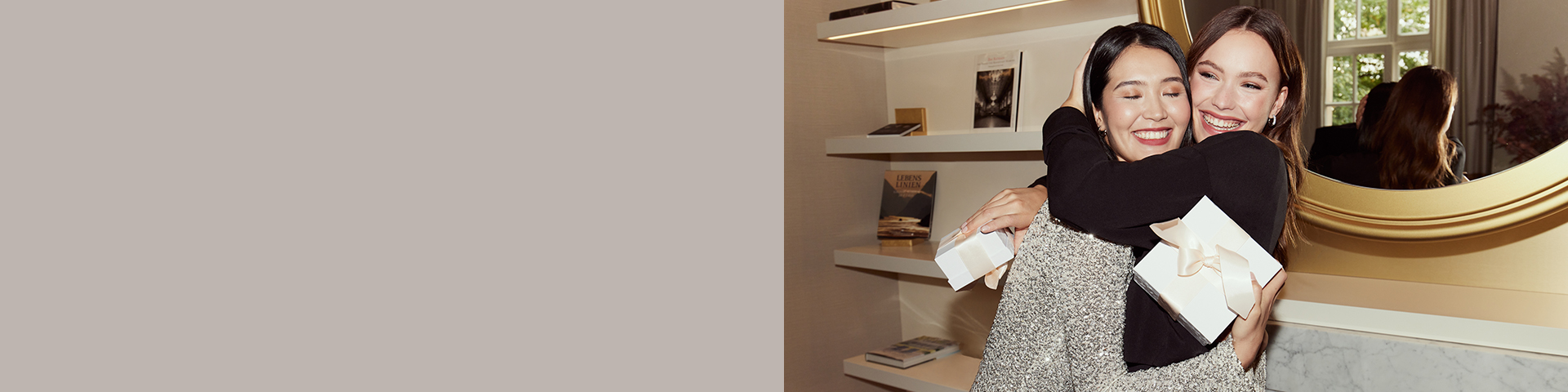 To kvinder i festligt t&oslash;j omfavner hinanden smilende og holder gave&aelig;sker i h&aelig;nderne. 