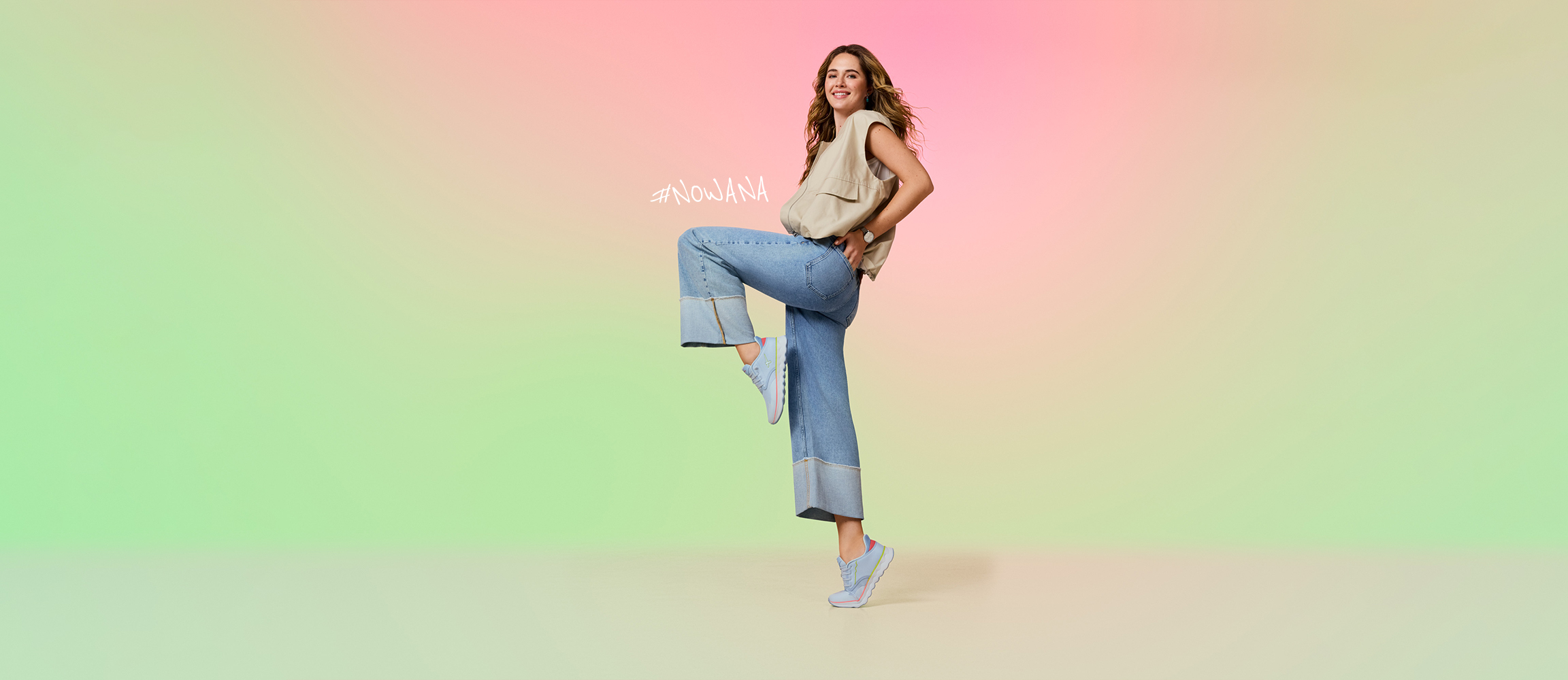 Woman in a beige top and jeans jumps happily in front of a pastel-colored background, her hair flowing in the air.