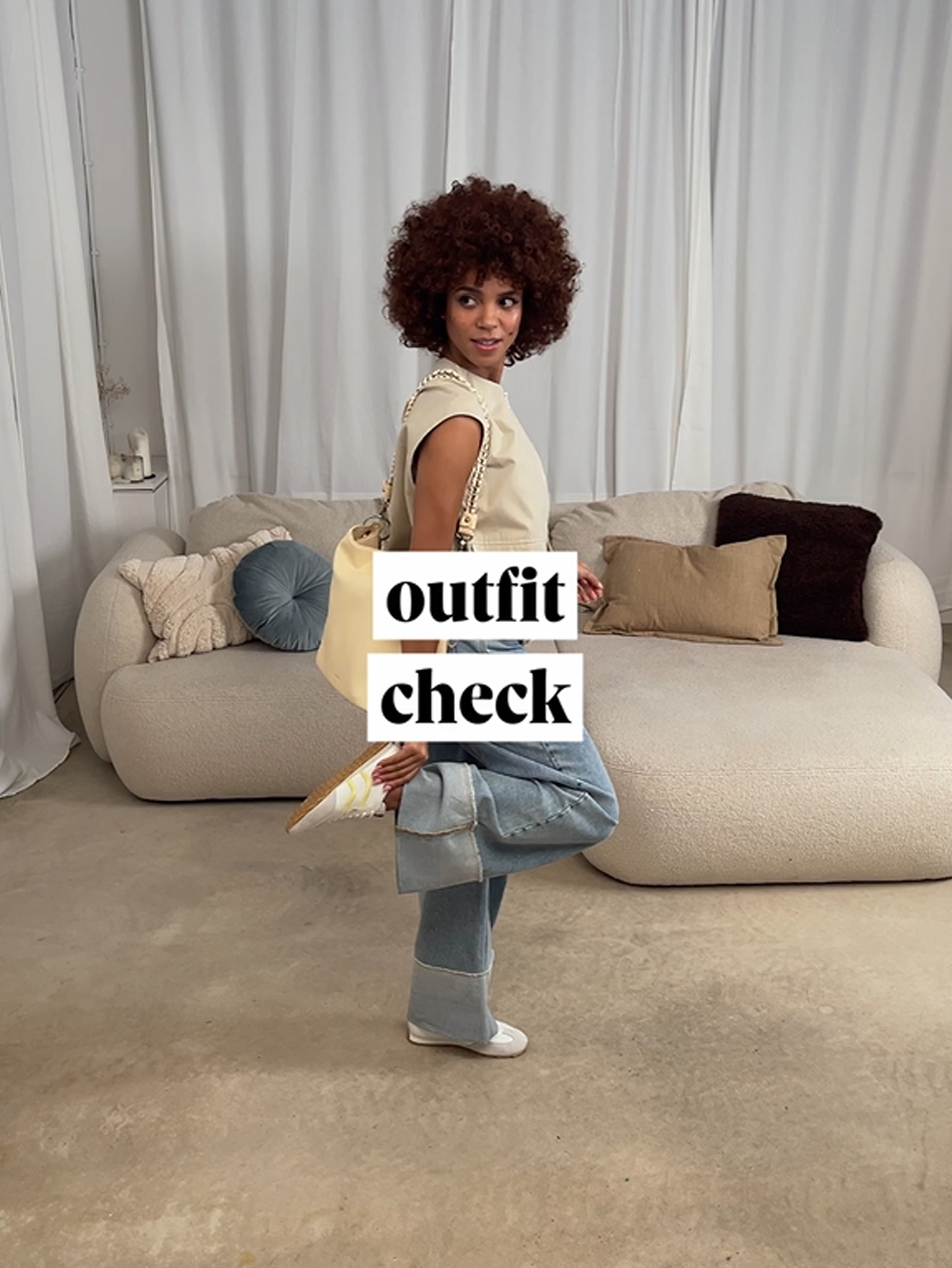 Frau mit lockigem Haar tr&auml;gt beige Weste, Jeans und helle Tasche, posiert vor einem cremefarbenen Sofa.