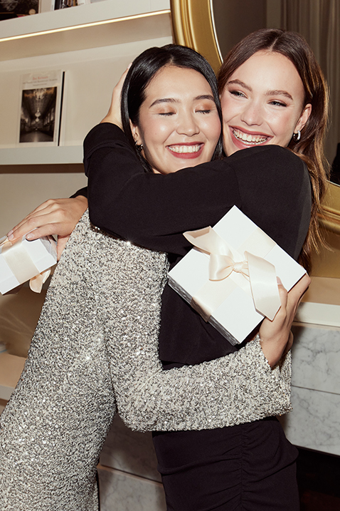 Two women in festive attire, one in silver sequins and one in black, embrace each other, laughing joyfully.