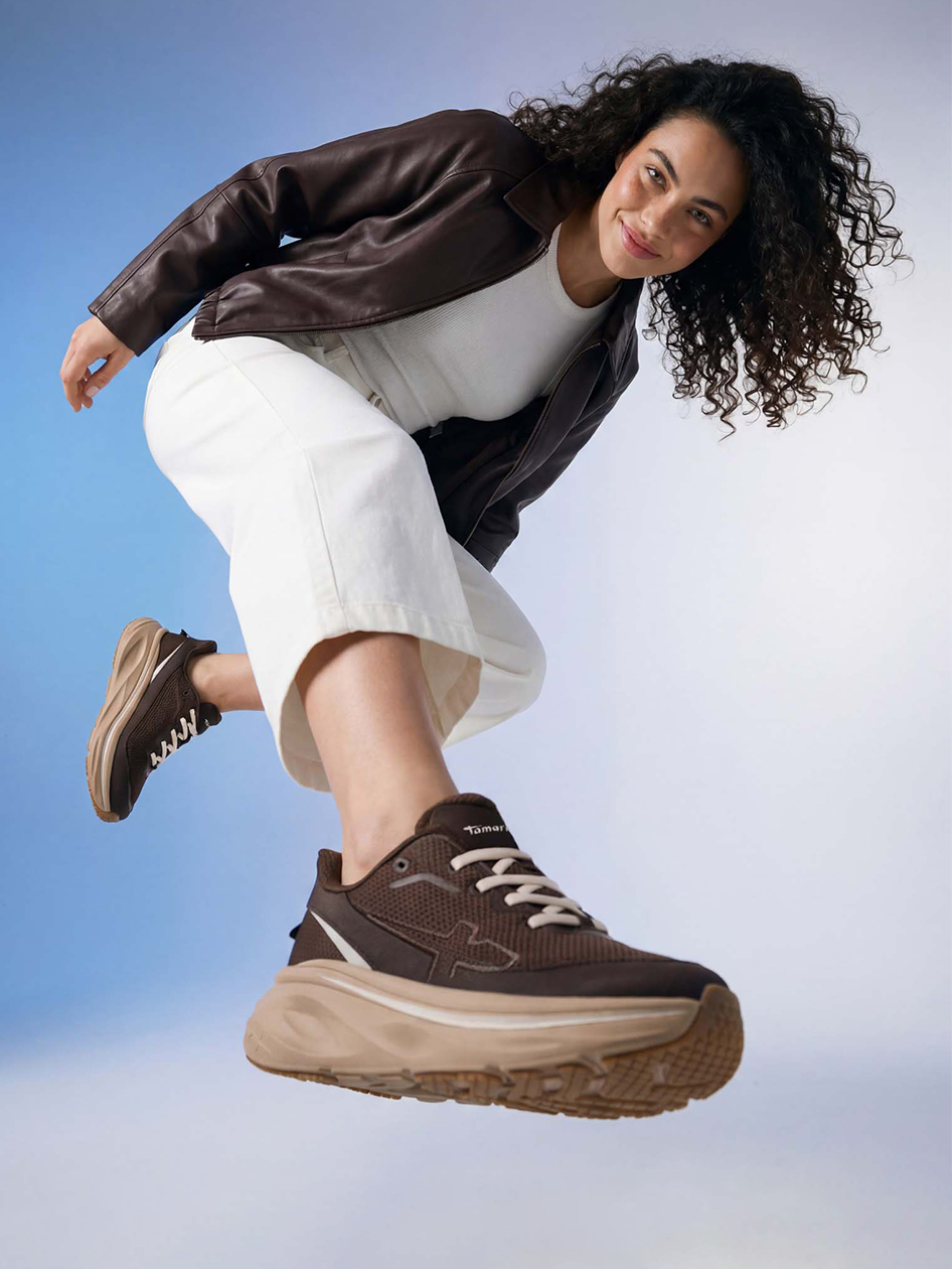 Kvinna med lockigt h&aring;r hoppar i brun skinnjacka, vita byxor och bruna Tamaris-sneakers framf&ouml;r en bl&aring; bakgrund.