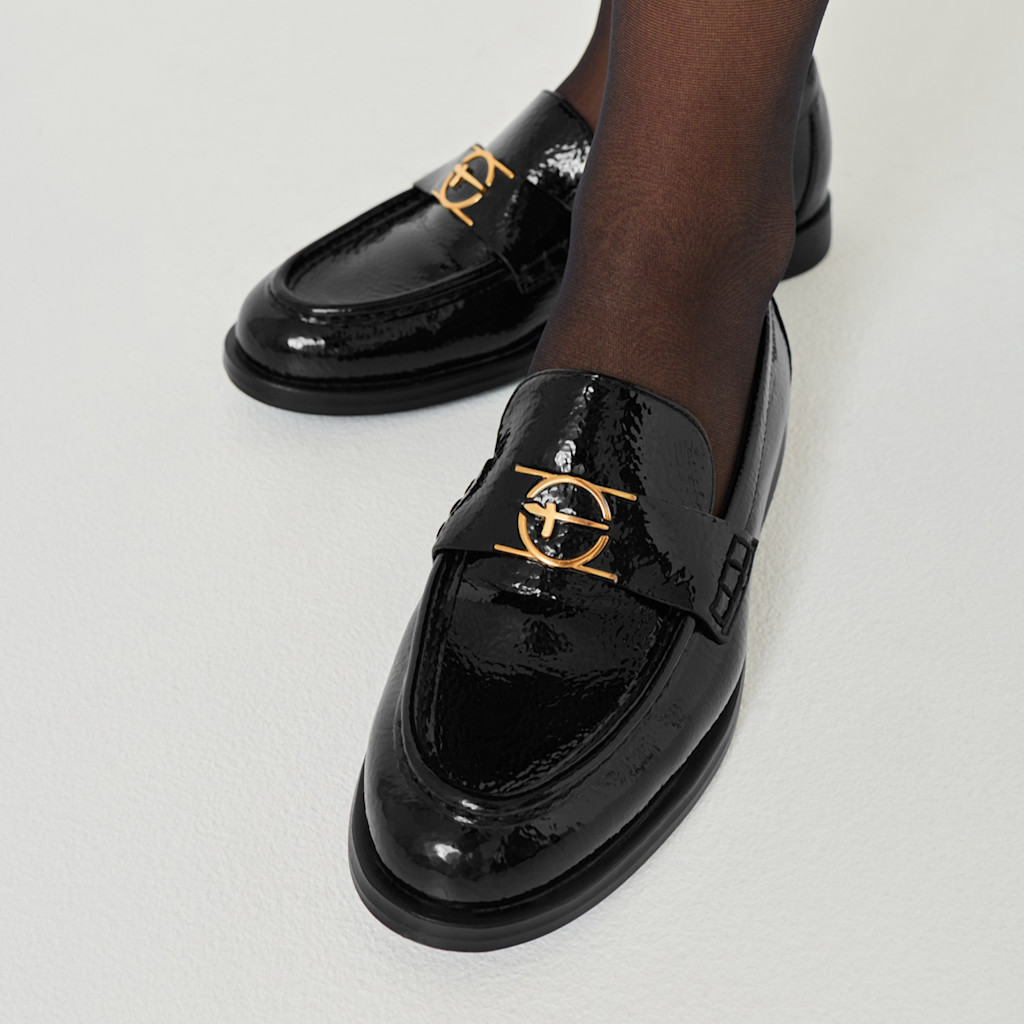 Shiny black patent leather loafers with gold metal hardware, worn with dark tights on a white background.