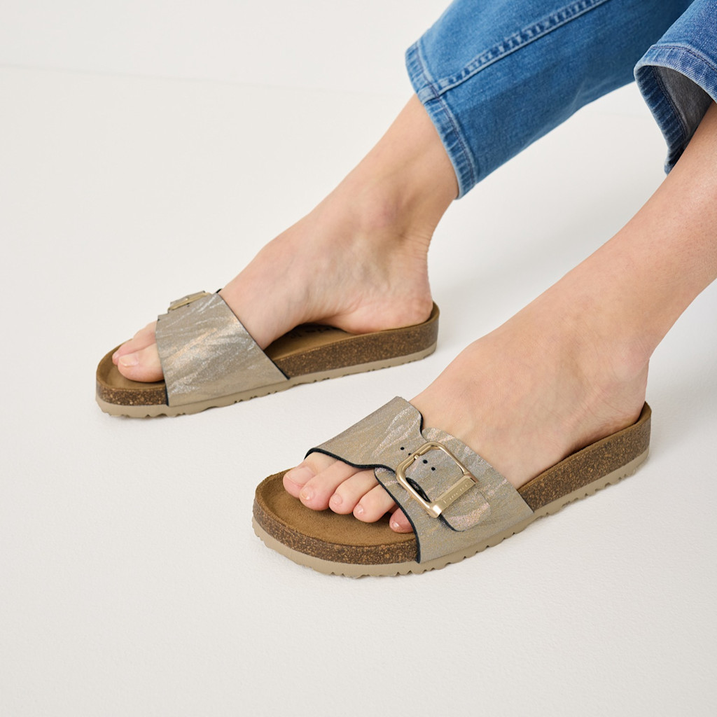 Goldbraune Pantoletten mit Schnalle und Korksohle, getragen zu einer blauen Jeans auf weißem Hintergrund.