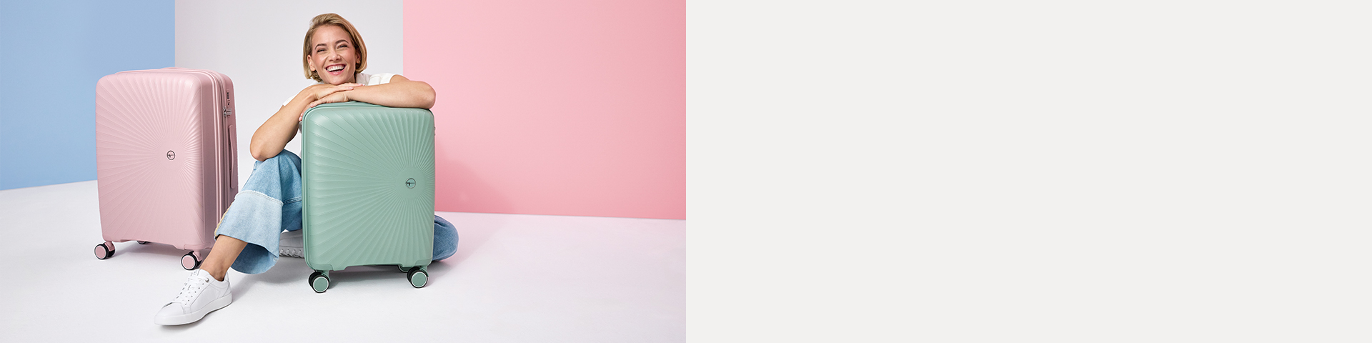 L&auml;chelnde Frau sitzt zwischen zwei Hartschalen-Koffern in Rosa und Mintgr&uuml;n vor pastellfarbenem Hintergrund.