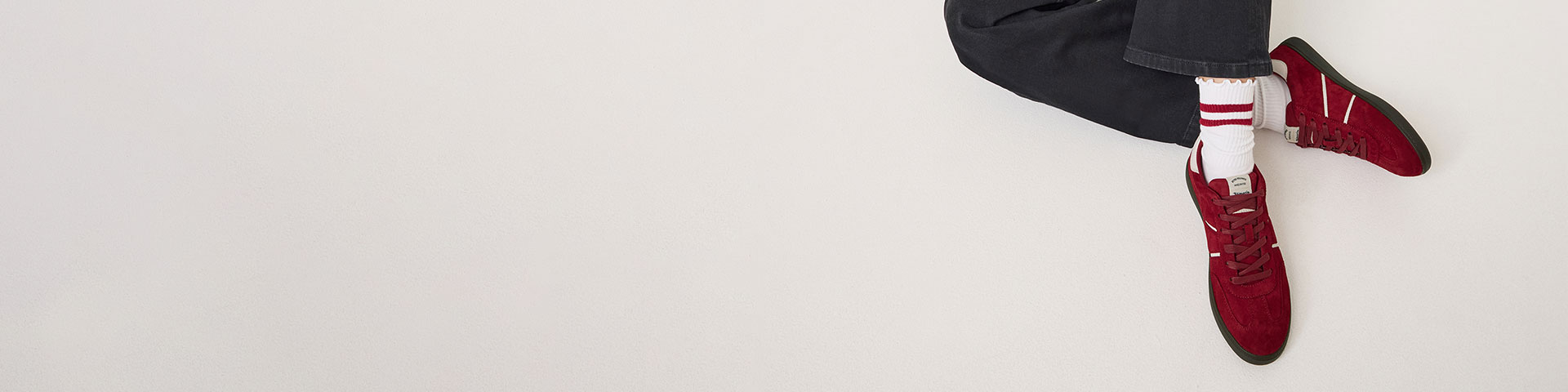 Füße in schwarzer Hose und roten Sneakers mit weißen Socken auf weißem Hintergrund, von oben fotografiert.