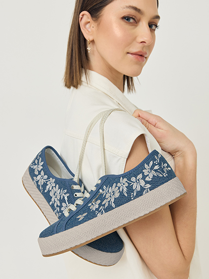 Vrouw houdt blauwe plateausneakers met witte bloemenborduursels in haar hand.