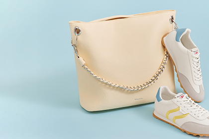 Borsa color giallo burro con dettaglio a catena accanto a sneakers bianche con strisce gialle su sfondo azzurro chiaro.