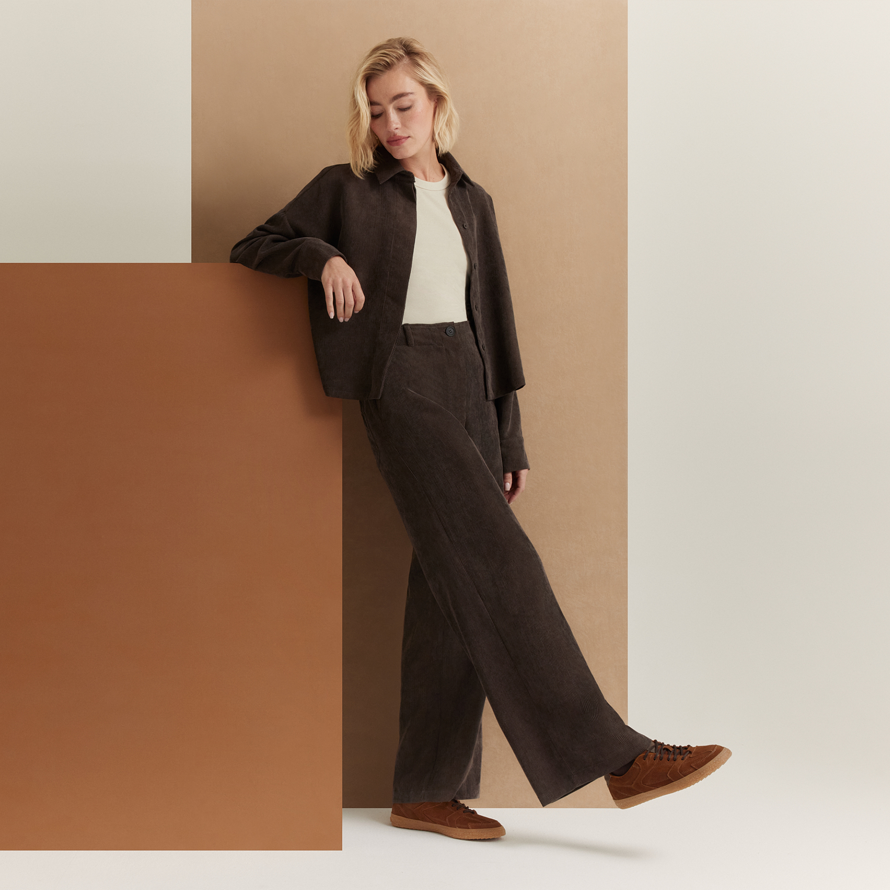 Žena v tmavě hněk&eacute;m man&scaron;estrov&eacute;m outfitu s b&iacute;l&yacute;m topem a hněd&yacute;mi teniskami se lež&eacute;rně op&iacute;r&aacute; o b&eacute;žovou zeď.