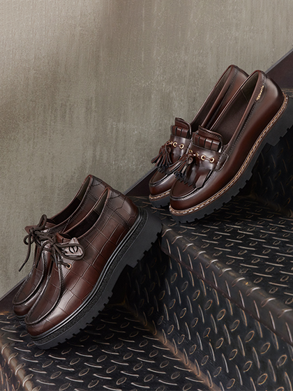 Zwei dunkelbraune Lederschuhe auf Metallstufen: ein Schn&uuml;rschuh mit Kroko-Muster und ein Loafer mit Quasten und dicker Sohle.