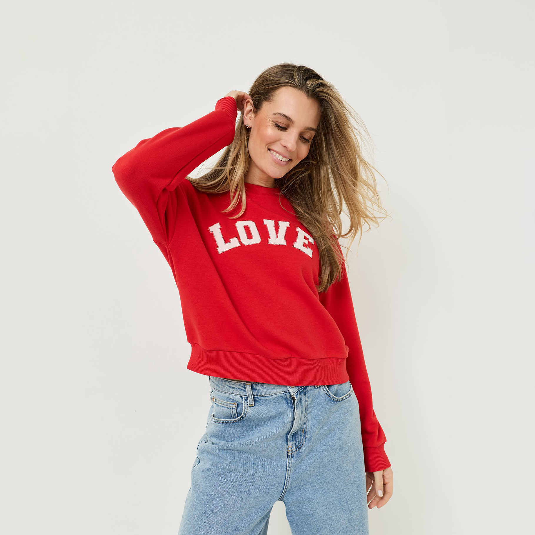 Frau mit langen Haaren tr&auml;gt rotes Sweatshirt mit wei&szlig;em "LOVE"-Schriftzug und hellblaue Jeans, l&auml;chelnd vor hellem Hintergrund.