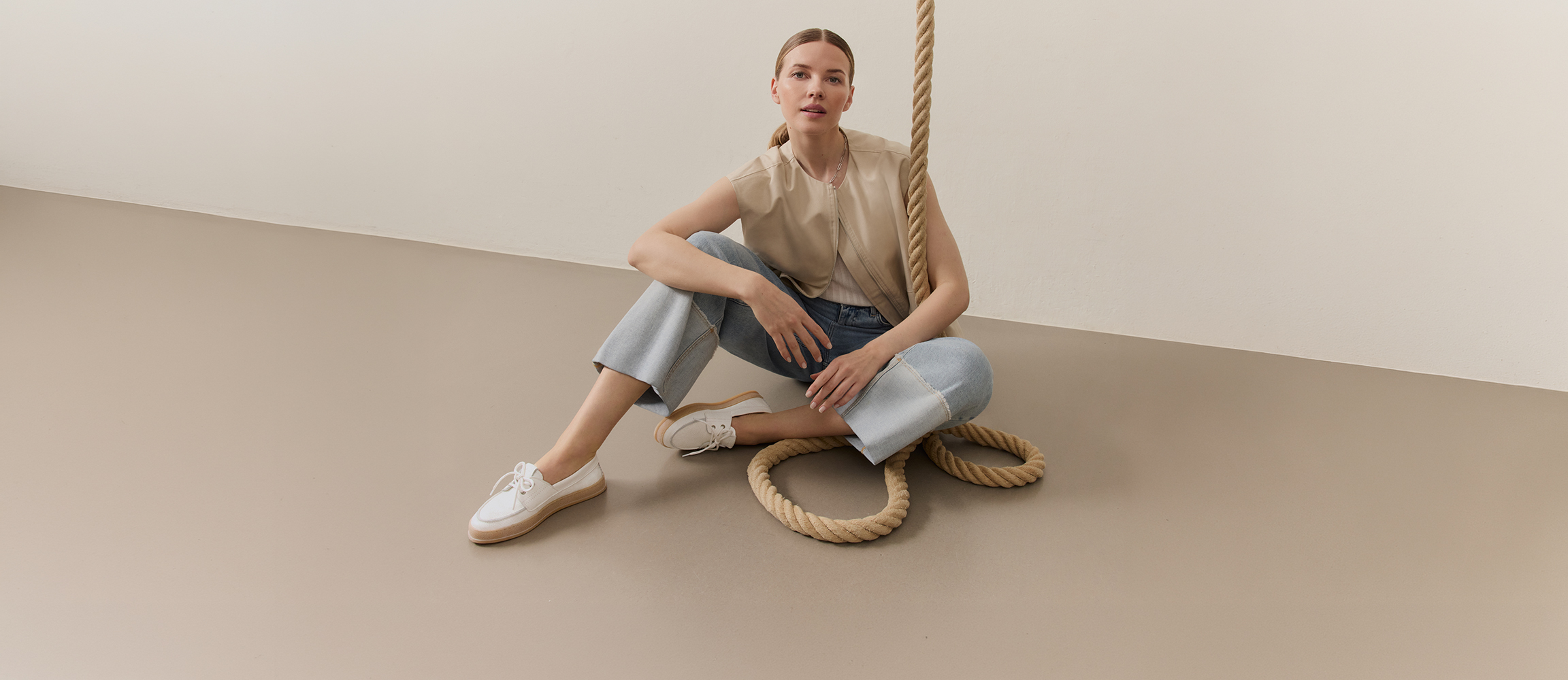Donna con top beige e jeans seduta in modo rilassato su un set minimalista con dettagli in corda, indossa boat shoes bianche.