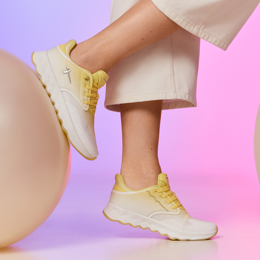 Chaussures de sport blanc et jaune port&eacute;es aux pieds sur fond rose-violet, associ&eacute;es &agrave; une tenue claire.
