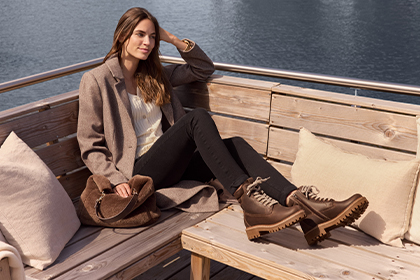 A woman is sitting relaxed on a wooden terrace in the sun, wearing warm autumn clothes and ankle boots.