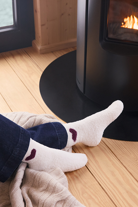 Feet in white socks with a dark red heart motif in front of a burning wood-burning stove on a wooden floor