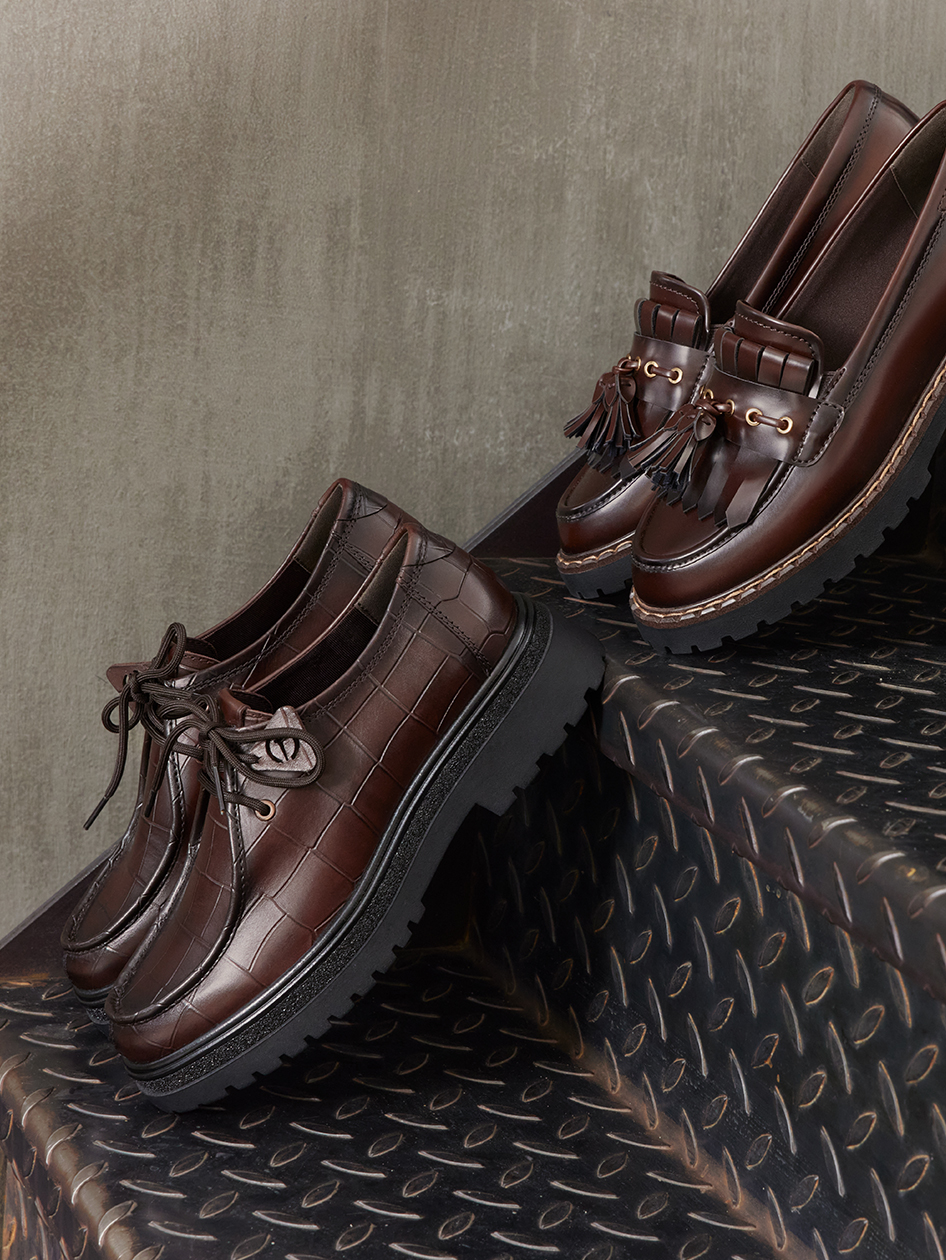 Twee donkerbruine leren schoenen: een veterschoen met krokoreli&euml;f en een loafer met kwastjes op een zwarte metalen trap.