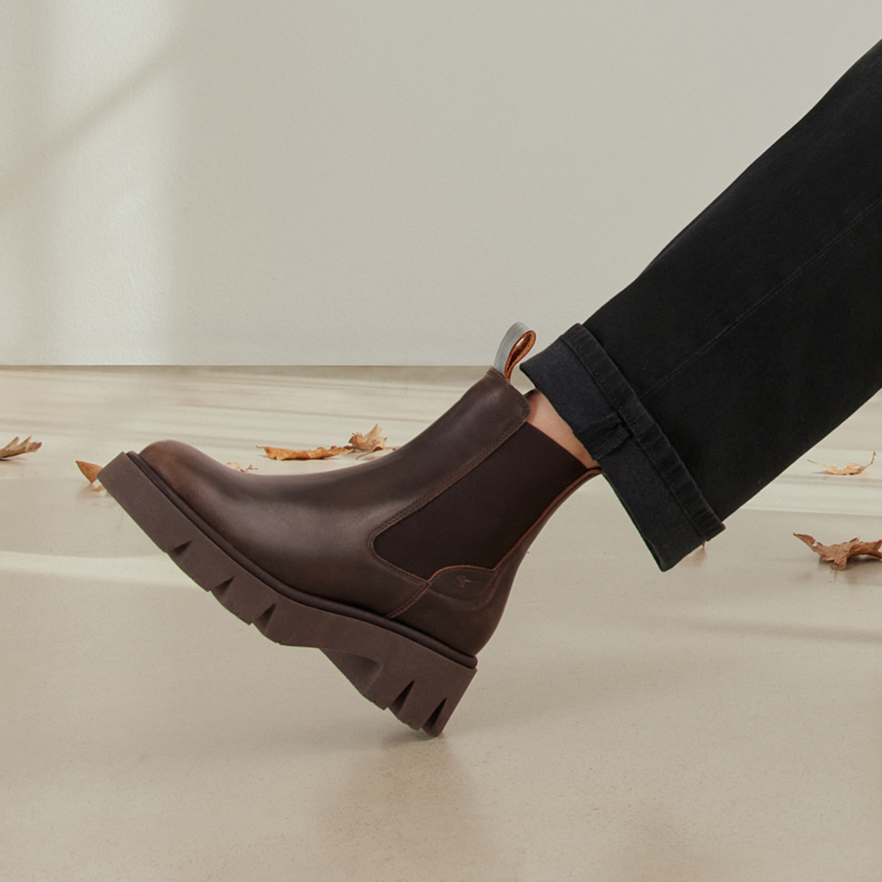 Boots Chelsea bruns avec semelle &eacute;paisse crant&eacute;e, port&eacute;s avec un jean noir, entour&eacute;s de feuilles d&rsquo;automne sur un sol clair.