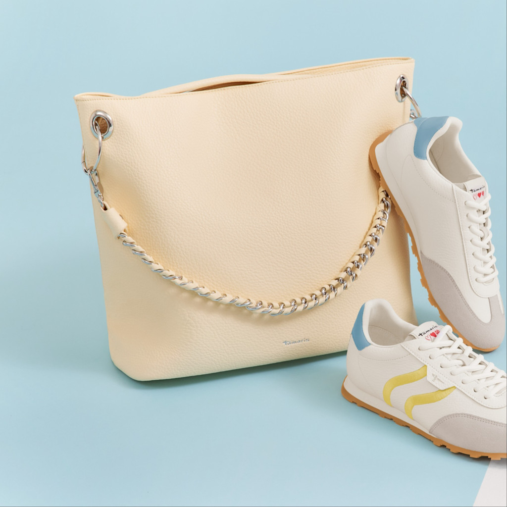 Cr&egrave;mekleurige handtas met kettingdetails naast witte sneakers met gele accenten op een lichtblauwe achtergrond