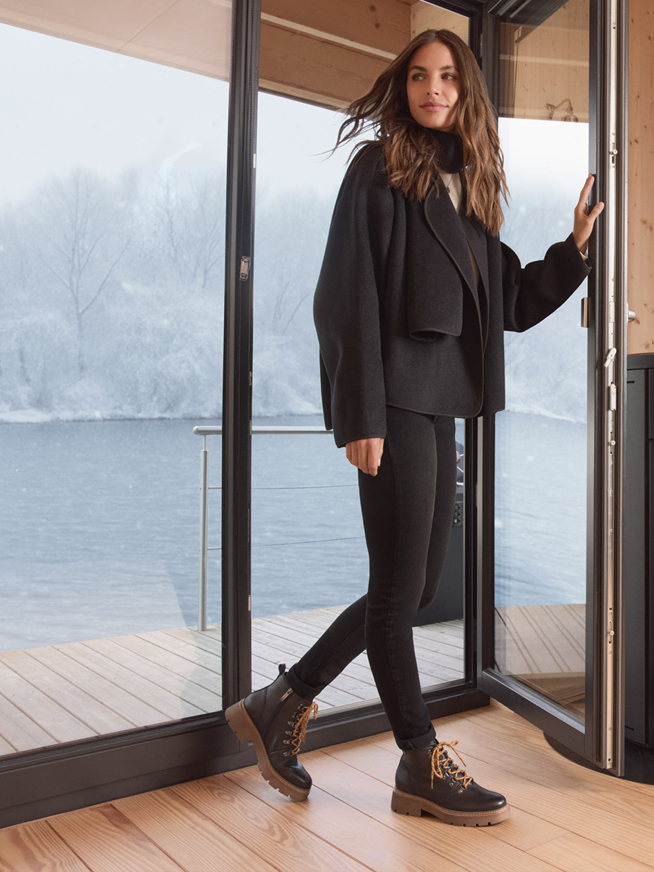 Une femme en tenue d'hiver noire et bottes de randonnée se tient devant des fenêtres avec vue sur le lac