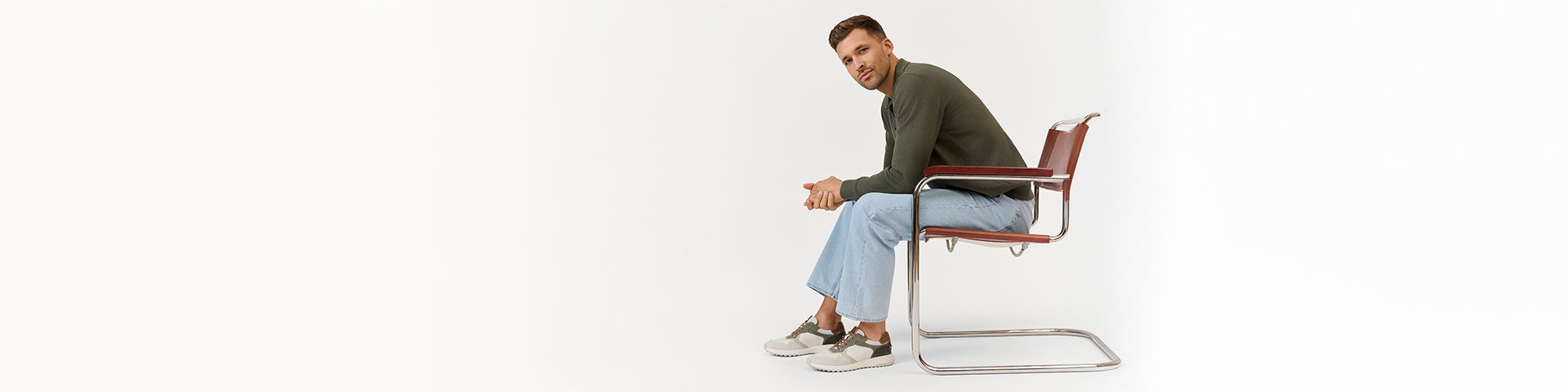 Persona con maglione verde oliva e jeans chiari seduta di lato su una sedia moderna con struttura in cromo.