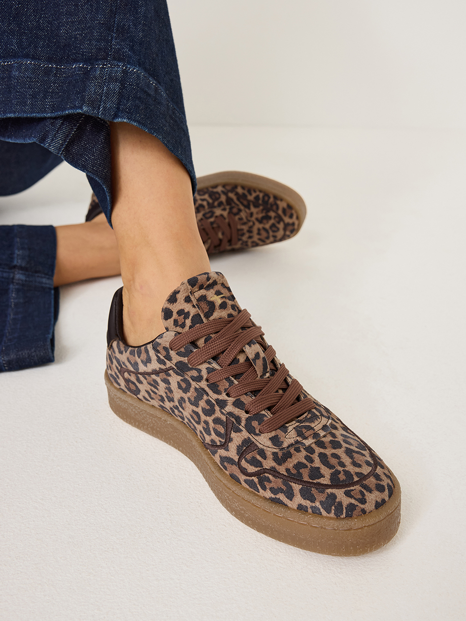 Sneakers met luipaardprint, bruine veters en rubberen zool, gedragen met donkere jeans tegen een witte achtergrond.