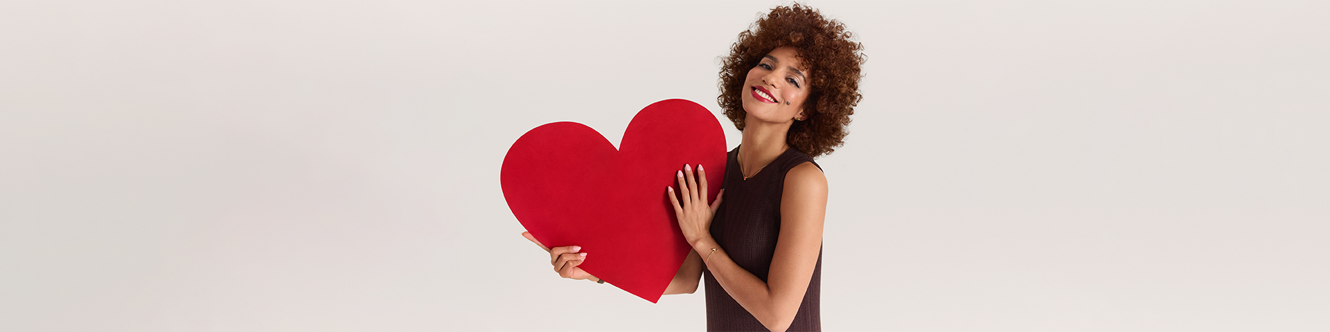 Smiling person with curly hair in a dark brown dress holding a large red heart against a white background