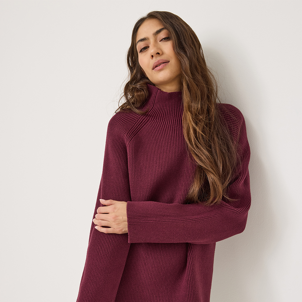 Giovane donna con maglione a collo alto bordeaux posa davanti a un muro bianco, porta i lunghi capelli castani a onde