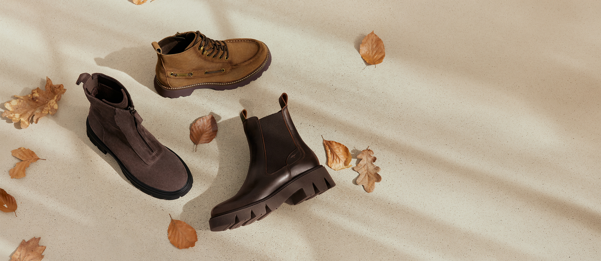 Three autumn ankle boots in brown and grey on a light surface with autumn leaves.